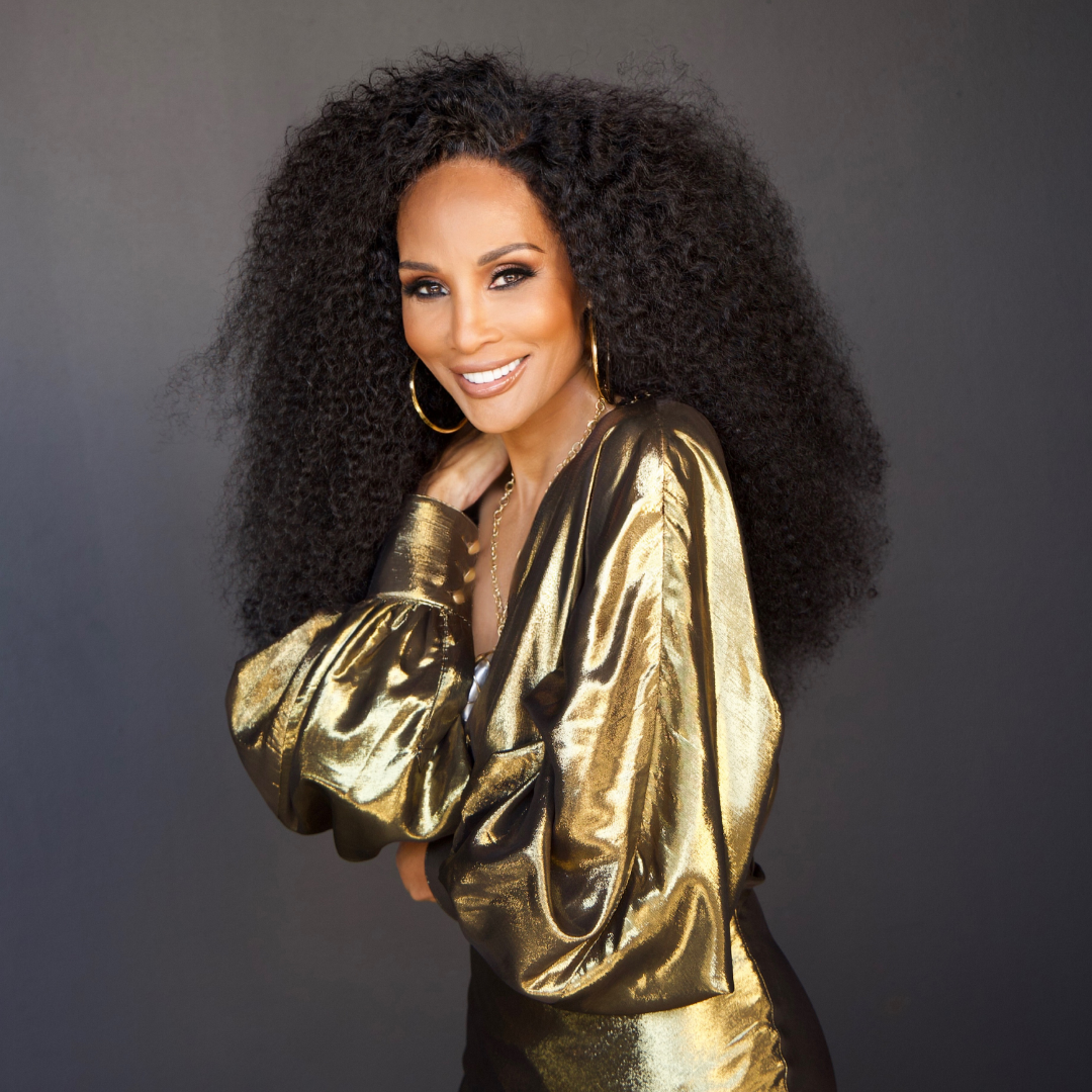 Person with long, curly hair in a shiny gold outfit, smiling against a neutral background.